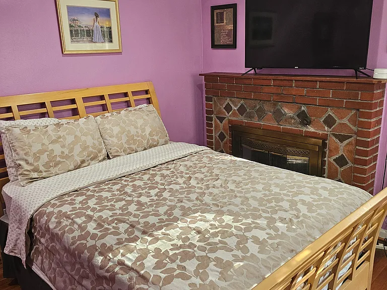 Furnished bedroom with a full bed, hardwood floors, and a brick fireplace mantel with TV for added convenience.