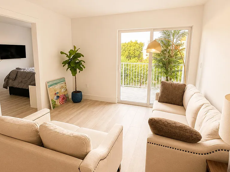 Open living area with comfortable seating, wall-mounted TV, and sliding doors leading to a balcony with leafy views.