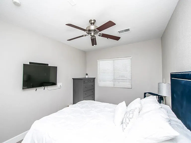 Casita bedroom suite at, Tempe rental home. Features a plush bed with crisp white bedding, a tufted navy velvet headboard, a wall-mounted flat-screen TV, a tall grey dresser, and a ceiling fan. Cozy, modern bedroom for a top Tempe, AZ stay.
