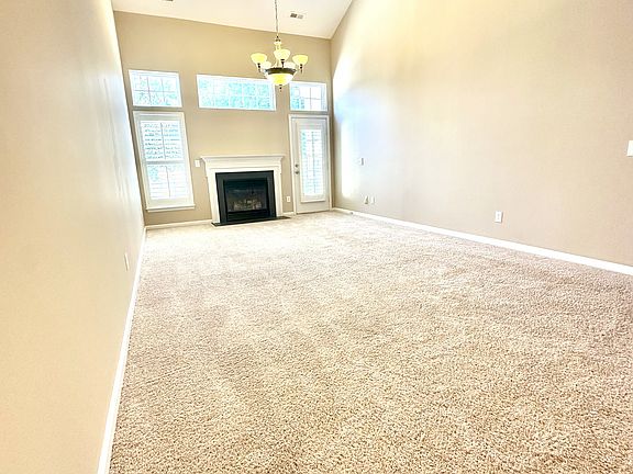 Spacious living room with vaulted ceilings, plush carpet, and a cozy gas fireplace. Tall windows and glass door fill the space with natural light_perfect for relaxing or entertaining