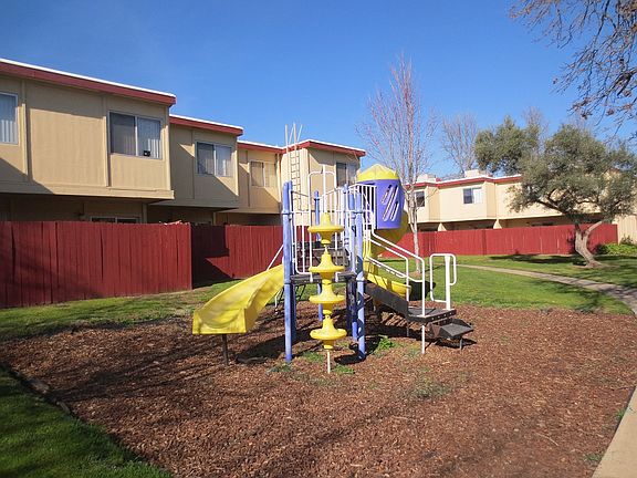 American River Gardens playground on bark with grass &amp; concrete surrounding, in front of apartment buildings with red fencing