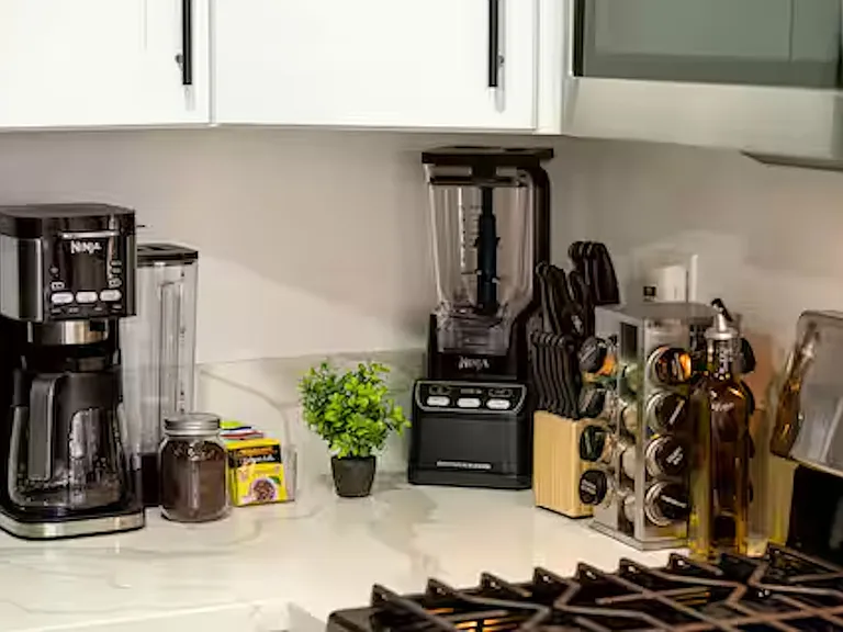 Countertop includes a spice rack, coffee and tea station, Ninja coffee maker and blender, with white stone benchtops and light timber flooring creating a fresh, welcoming feel.
