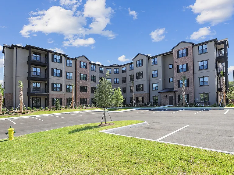 Modern four-story apartment building with brick and stucco facade, surrounded by lush landscaping and ample parking, under a clear blue sky.