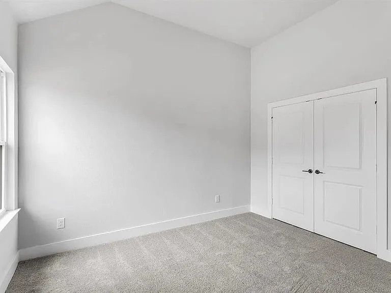 Spacious room featuring a vaulted ceiling, light gray carpet flooring, and a large window with white trim