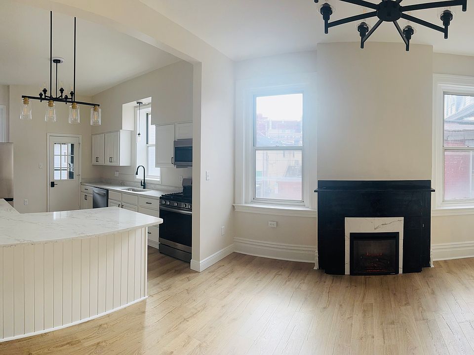 Bright, open-concept living space with a modern kitchen featuring quartz countertops, stainless steel appliances, and an island with seating. The sunlit living area offers warm wood floors and a sleek fireplace, perfect for relaxing or entertaining.