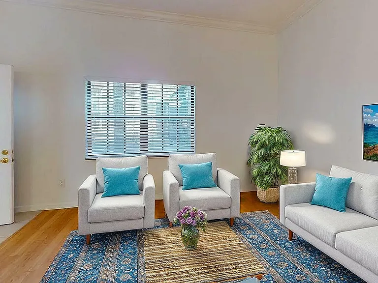 Inviting living room with stylish white sofas, blue accent pillows, and a vibrant area rug, creating a cozy atmosphere. Large window with blinds and wall-mounted artwork enhance the bright and airy space.