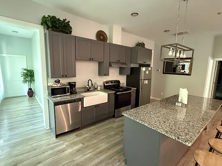 Open kitchen with ample gray cabinetry, granite-look counters, farmhouse sink, and stainless appliances plus a prep island.