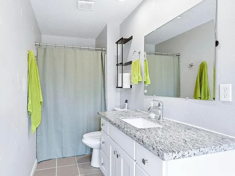Full bath with granite-top vanity, large wall mirror, and shower/tub combo with curtain, plus open shelving for storage.