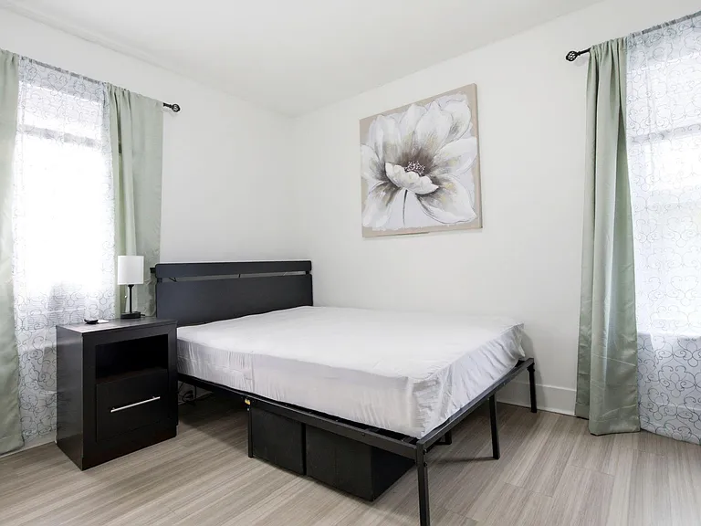 Furnished bedroom with a full-size bed, nightstand, and dual windows with curtains for privacy and natural light.