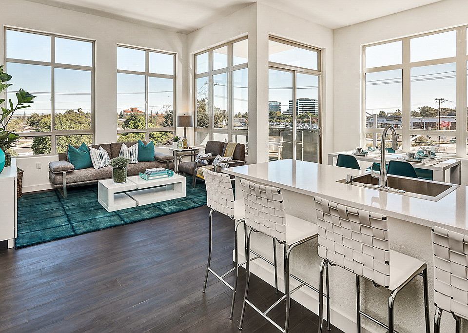 Staged, open living room and kitchen with hardwood-style, vinyl flooring and floor to ceiling windows.