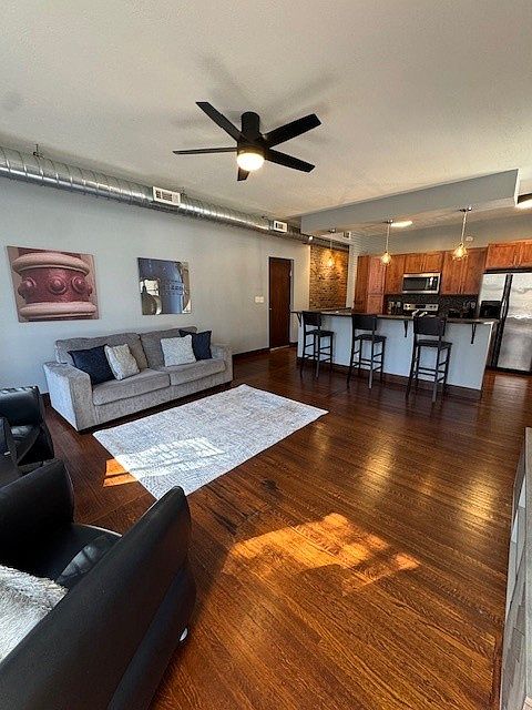 Stylish open-concept living area with a gray sofa, cozy rug, and sleek black chair. The kitchen features warm wood cabinets, stainless steel appliances, and a countertop with barstools. Dark wood floors and exposed ductwork add an industrial-modern vibe.
