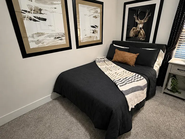 Well-appointed bedroom featuring a full bed setup, soft carpet, and a convenient nightstand beside a window with blinds.