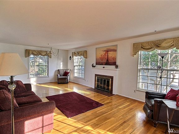 A spacious living room with brick wood-burning fireplace that offers a white solid wood mantel and there are 4 large bright windows on each exterior wall.