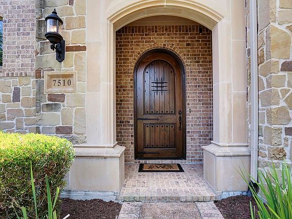 This welcoming front porch is accentuated by a gorgeous stucco columned archway that leads to a beautiful solid wood, arched front door. Notice the brick paved porch floor as well as the Mediterranean styled outdoor sconce light.