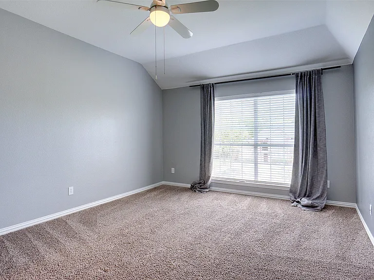 Spacious room featuring vaulted ceilings, neutral-toned carpeting, a ceiling fan with integrated lighting, and a large window with blinds and drapes