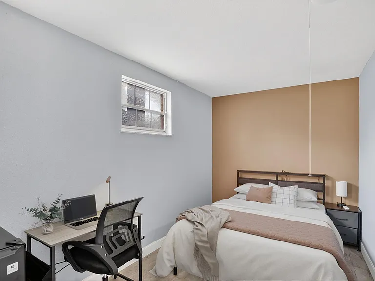 Furnished bedroom with a full bed, nightstand, and a dedicated desk area, plus a window for natural light.