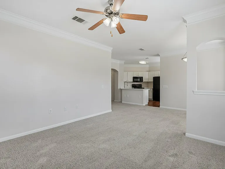 Inviting open-plan living area with plush carpeting, a modern ceiling fan, and a sleek kitchen featuring white cabinetry and black appliances. Inviting open-plan living area with plush carpeting, a modern ceiling fan, and a sleek kitchen featuring white c