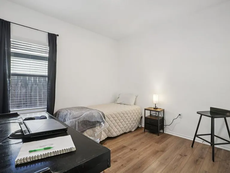 Private bedroom with wood-look flooring, a window with blinds, and space for a desk, plus a nightstand and lamp.
