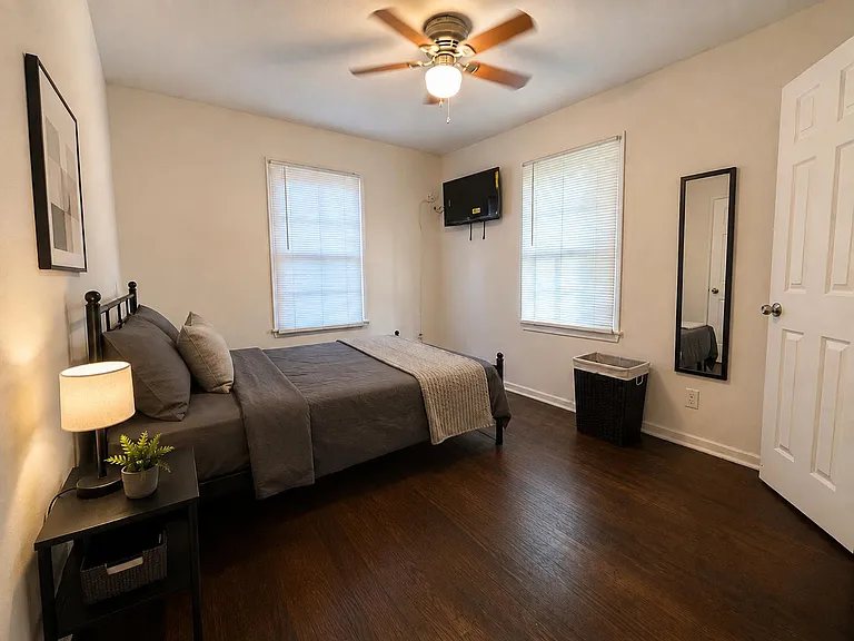 Furnished bedroom with hardwood floors, ceiling fan, wall-mounted TV, and two windows with blinds for natural light.