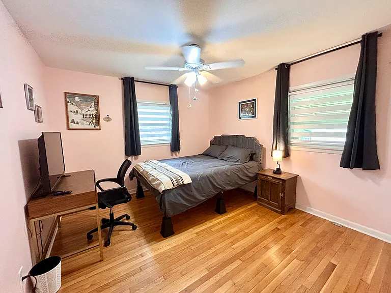 Furnished bedroom with hardwood floors, dual windows with treatments, ceiling fan, and a desk/TV setup for work and relaxing.