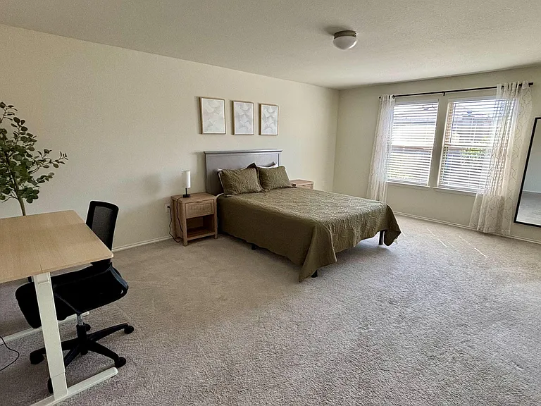 Spacious carpeted bedroom with a large window, queen bed, bedside table, full-length mirror, and a dedicated desk area.