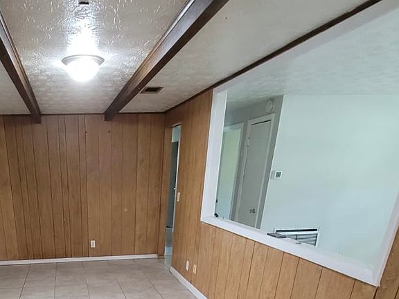 Open dining area flowing seamlessly into the kitchen, featuring wood-and-gray accent walls, a white ceiling with bright lights, and two long wooden beams for a modern, stylish touch.