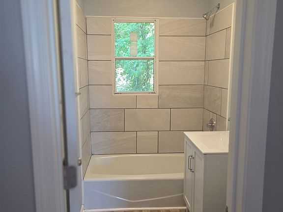 Beautifully renovated full bathroom featuring a modern tiled tub and shower surround, updated vanity, and durable wood-style flooring. A large window provides natural light while enhancing the bright, clean atmosphere. Contemporary finishes and neutral to