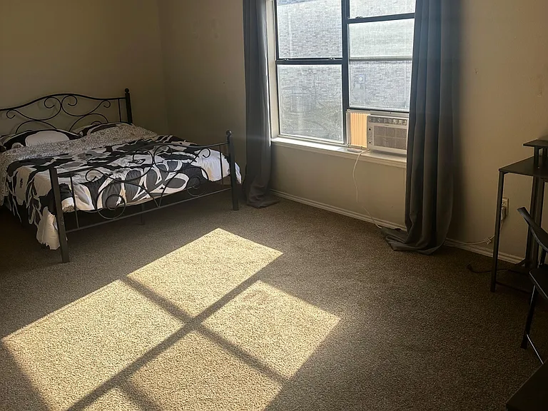 Carpeted bedroom with a large window, blackout curtains, and window A/C, plus space for a desk or dresser.