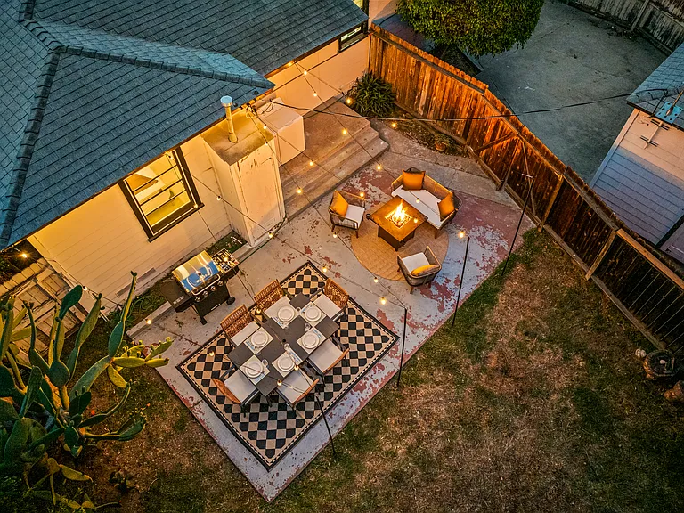 Cozy backyard retreat with cafe-style string lights, outdoor dining, a BBQ grill, and a fire pit lounge. Surrounded by palm trees and warm lighting, this inviting space is perfect for relaxing evenings and group gatherings.
