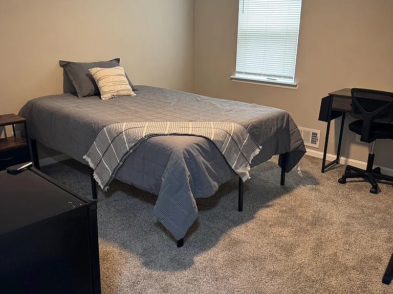 Private bedroom with a furnished bed, carpeted floors, ceiling fan, and a dedicated desk area near the window.