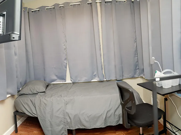 Compact furnished bedroom with single bed, desk/work area, and window wall with curtains for added privacy.