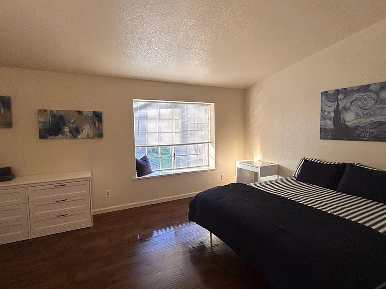 Spacious bedroom with hardwood floors, a large window with blinds, and room for a full bed plus dresser or desk.