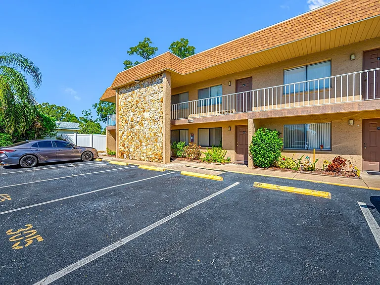 Charming two-story apartment building with a stone facade, surrounded by lush greenery and ample parking space, under a clear blue sky.