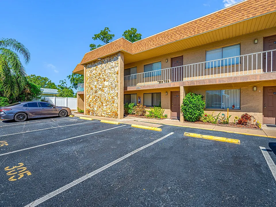 Charming two-story apartment building with a stone facade, surrounded by lush greenery and ample parking space, under a clear blue sky.