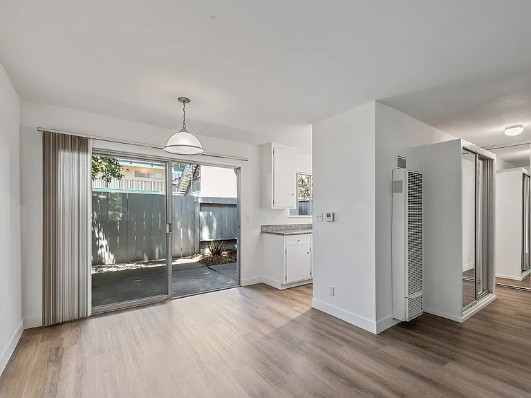 A view from the living room, showing the patio doors leading to a private outdoor space, the dining area and kitchen, as well as the hallway leading to the bedroom and bathroom.