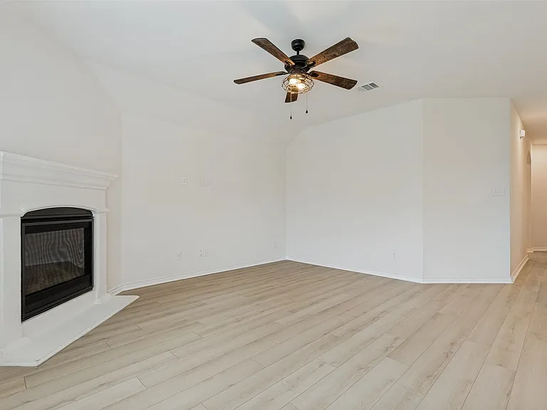 Spacious interior featuring a built-in fireplace with a white mantel, light wood-finish flooring, and a ceiling fan with integrated lighting