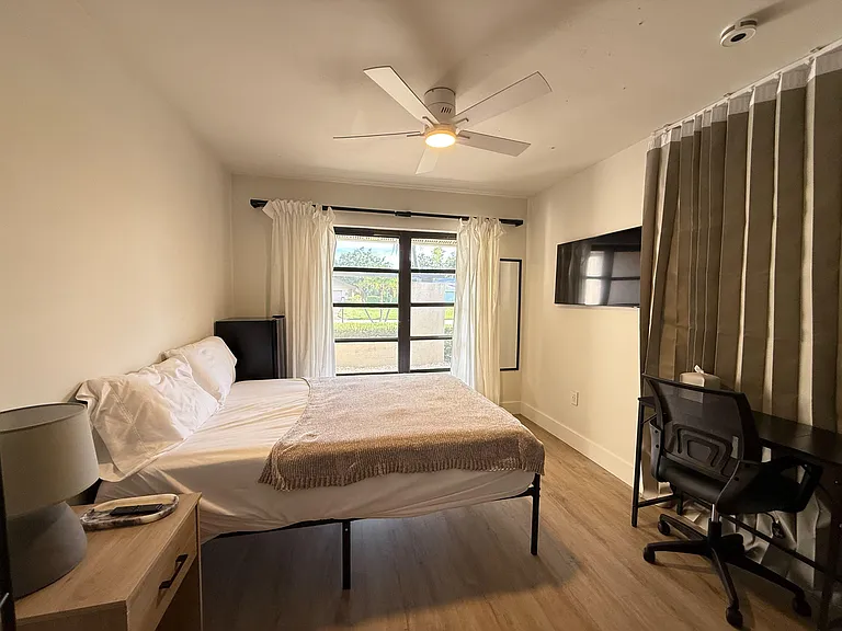 Furnished bedroom with a full bed, ceiling fan, and a wall-mounted TV, plus a work desk and large window with curtains.
