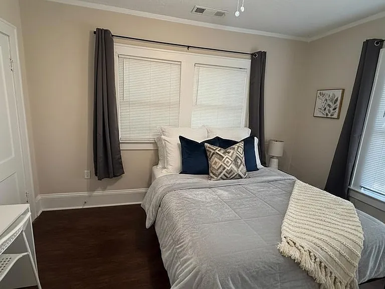 Well-appointed bedroom with a comfortable bed, closet, desk, and two windows with blinds and curtain panels.