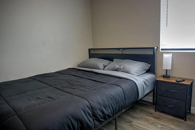 Furnished bedroom with a queen bed, nightstand with lamp, and window blinds for light control over durable flooring.