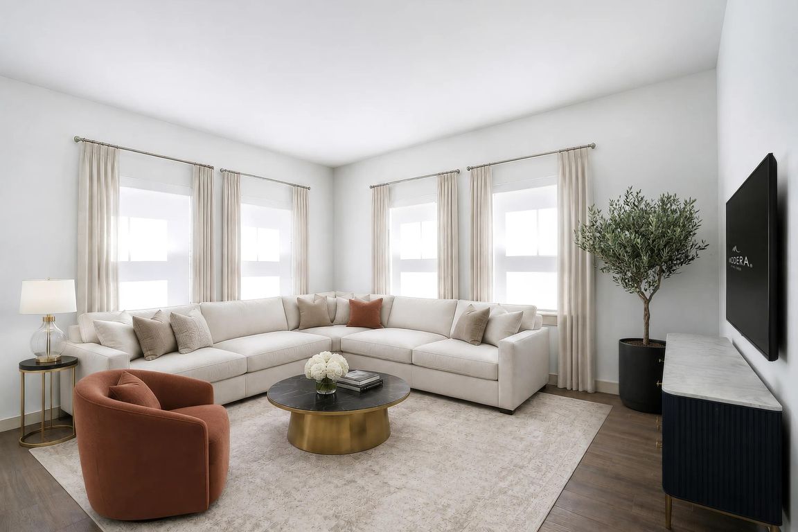 Light-filled living room with expansive corner windows, warm flooring, and ample seating designed for gathering, relaxing, and everyday comfort.