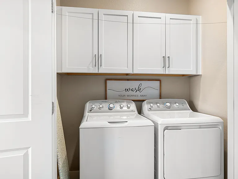 Clean and functional laundry room featuring a white washer and dryer set, complemented by white overhead cabinets for ample storage. A charming sign that reads "wash your worries away" adds a cozy, welcoming touch to this practical space.