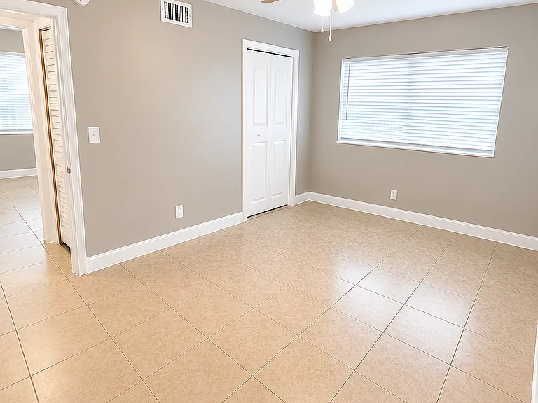 Bright, spacious bedroom featuring elegant tile flooring, a large window with blinds, and a ceiling fan.