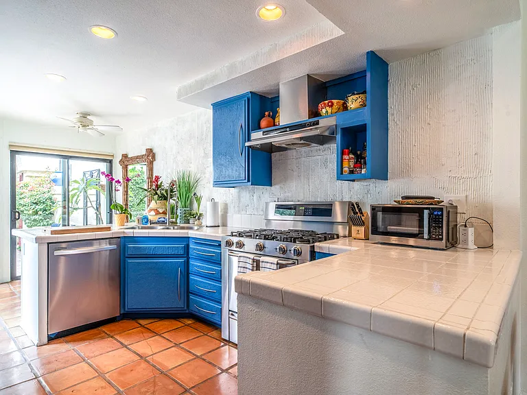 Bright, fully equipped kitchen featuring custom blue cabinetry, stainless gas range, tile countertops, and natural light throughout. Designed for extended stays with quality cookware and seamless access to the courtyard.