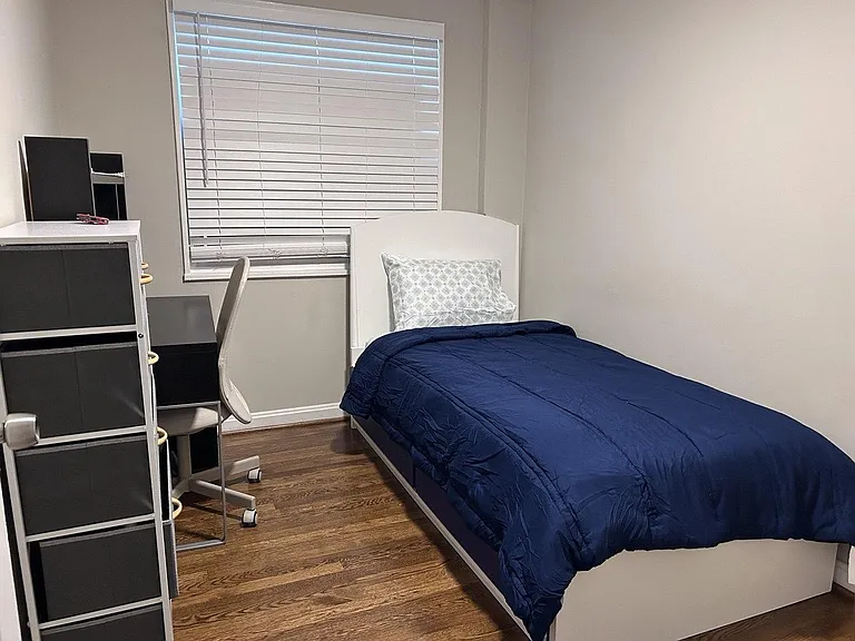 Furnished bedroom with a single bed, work desk and chair, and a window with blinds over hardwood-style flooring.