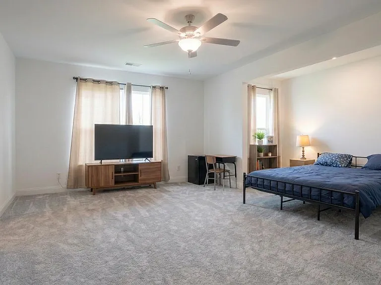 Spacious bedroom with plush carpet, ceiling fan, large windows, and a work nook plus TV stand and shelving.