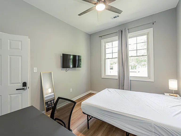 Private bedroom with a large window, ceiling fan, wall-mounted TV, and space for a bed and desk setup.