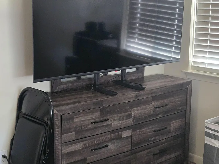 Bedroom includes a dresser with TV setup, carpeted floors, and a window with blinds for adjustable natural light.