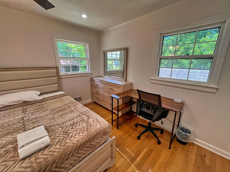Furnished bedroom with hardwood floors, two large windows, dresser with mirror, and a dedicated desk workspace.