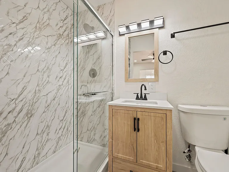 Renovated bathroom with a glass-enclosed shower, marble-look surround, and a vanity with storage and updated lighting.