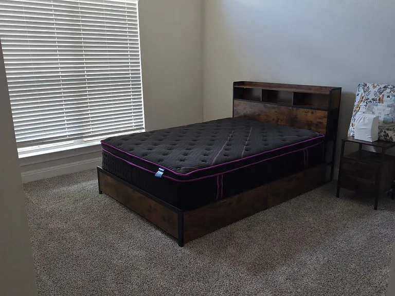 Carpeted bedroom with a high window and blinds, offering a comfortable layout for a full/queen bed and nightstand.